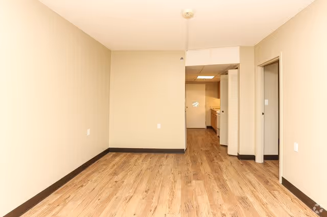 Empty room with light beige walls and wood flooring, showing an open doorway leading to a small kitchen area with cabinets and appliances, and another doorway to the right.