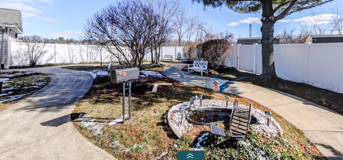 Outdoor garden area with a small pond surrounded by white rocks and a miniature wooden bridge. There is a mailbox on a post, a white bench, and a curved concrete walkway. Leafless trees and bushes are visible, along with a white privacy fence enclosing the area. The sky is clear with a few clouds.