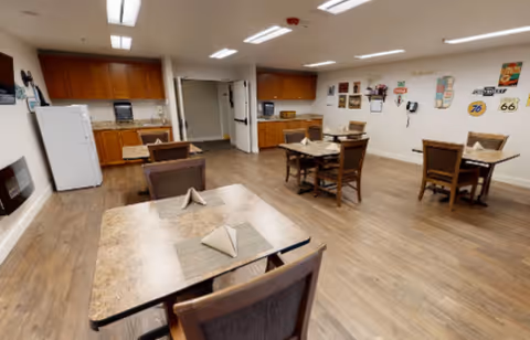 A dining area in Peters Creek Retirement Center with several tables and chairs arranged on a wooden floor. Each table has placemats and folded napkins. The room has a kitchenette with wooden cabinets, a refrigerator, microwave, and a coffee maker. The walls are decorated with vintage-style signs and posters.