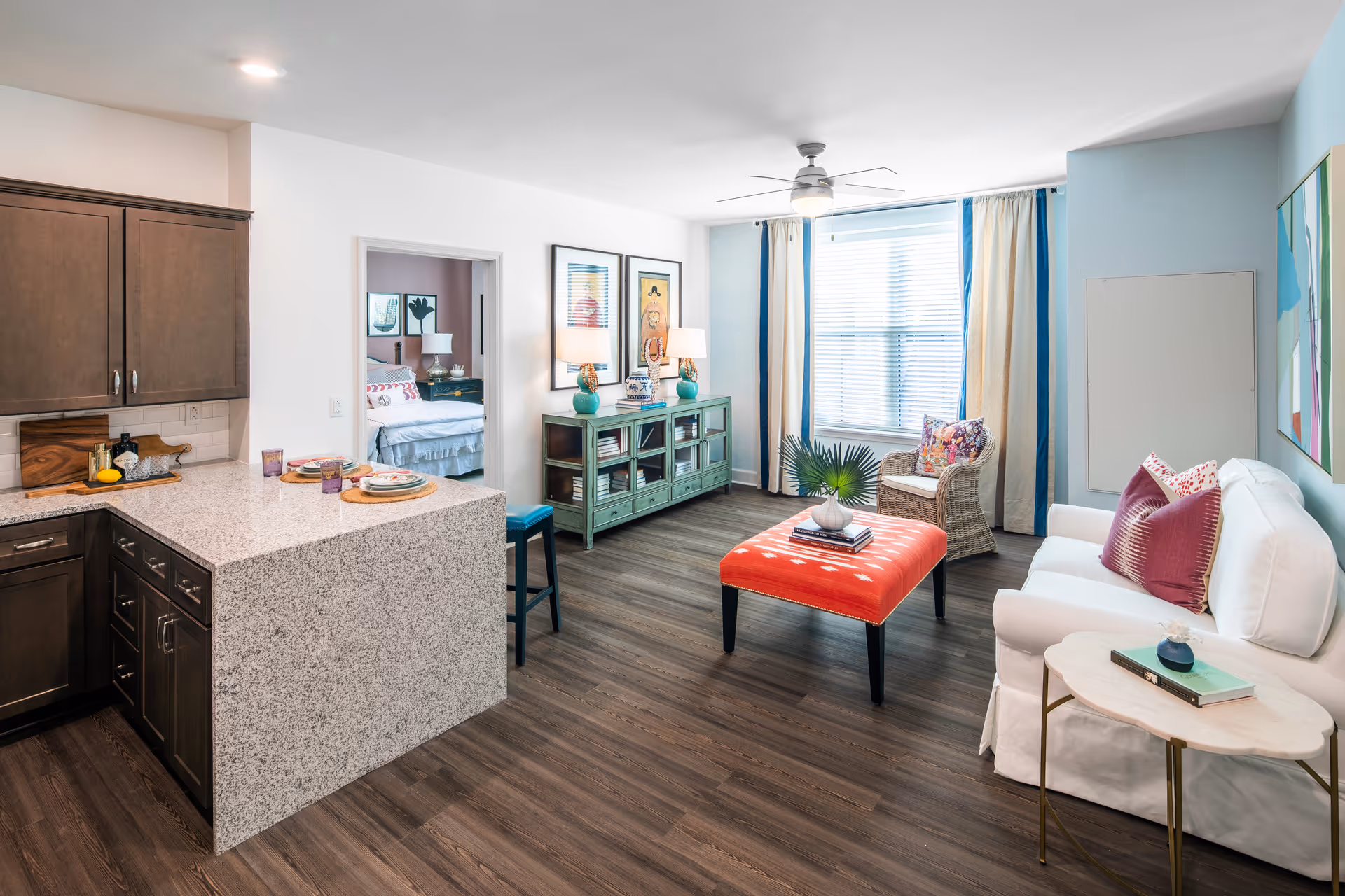 Bright open-plan living room and kitchen with a white sofa, red ottoman, granite island, and a view into a bedroom.