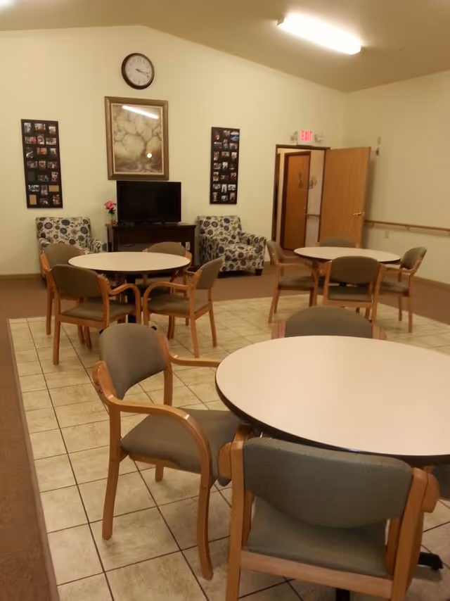 A common area in an assisted living facility with round tables and chairs arranged on a tiled floor. Two patterned armchairs are placed against the far wall near a TV on a wooden stand. Above the TV is a framed picture and a clock. The walls are light-colored, and there is an open door leading to another room.