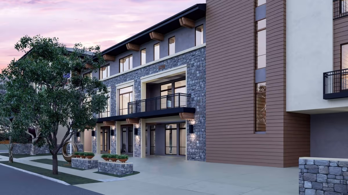 Front exterior of a modern multi-story senior living building with stone and wood siding, balconies, landscaping, and a paved entrance.