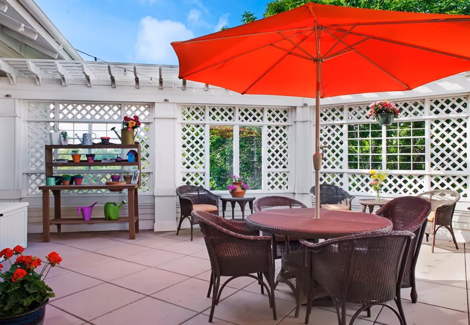 Outdoor patio area with a round table and six wicker chairs under a large red umbrella. The patio is enclosed with white lattice walls and decorated with potted plants and flowers on shelves and tables. The sky is blue with some clouds visible above.