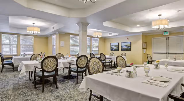 A dining room in a senior living facility with multiple tables covered in white tablecloths, each set with plates, glasses, and silverware. The room has patterned chairs, large windows letting in natural light, yellow walls, and ceiling light fixtures.