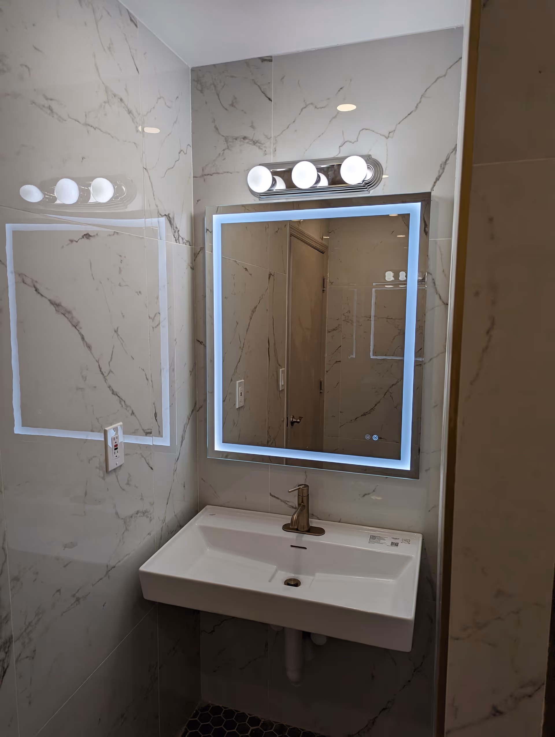 A modern bathroom sink area with a rectangular white sink mounted on a marble-patterned wall. Above the sink is a square mirror with built-in LED lighting around the edges. Above the mirror is a light fixture with three round bulbs. The reflection in the mirror shows a door and part of the bathroom.