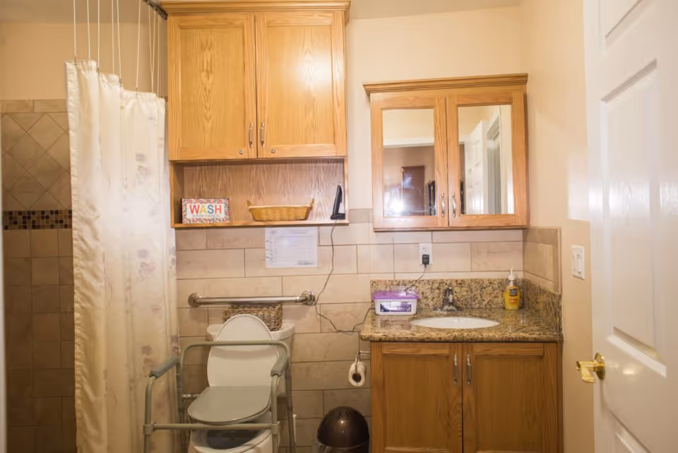 A bathroom with beige tiled walls and a shower area with a floral shower curtain on the left. There is a toilet with a safety frame around it and a grab bar on the wall behind. Above the toilet, there is a wooden cabinet and a small shelf holding a decorative sign that says 'WASH your hands' and a basket. To the right, there is a granite countertop with a sink, a soap dispenser, and a purple container. Above the sink is a wooden medicine cabinet with mirrored doors. A white door with a gold handle is partially visible on the right side.