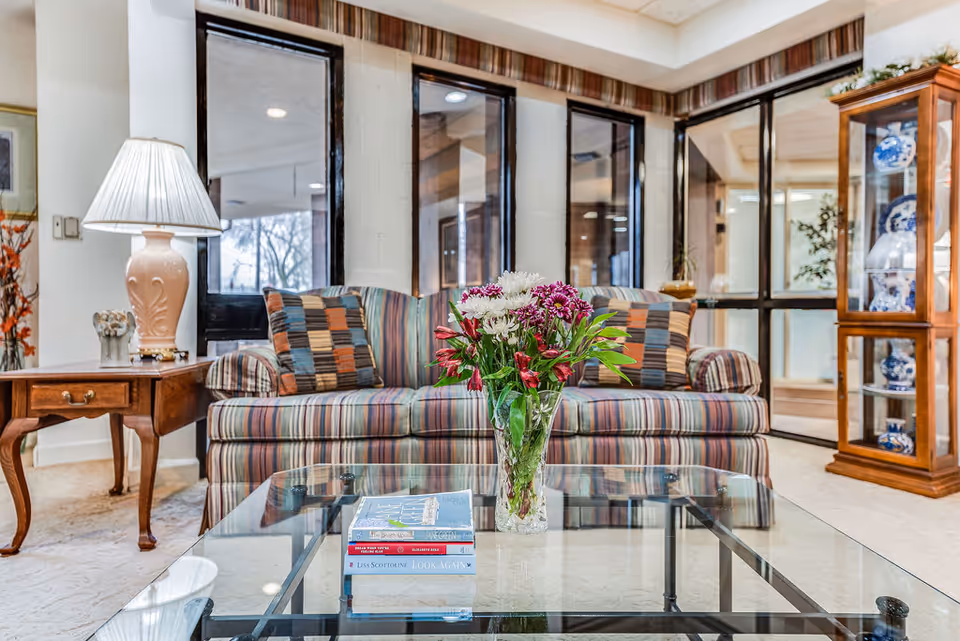 A cozy living room area featuring a striped sofa with two patterned cushions, a glass coffee table with a vase of fresh flowers and a stack of books, a wooden side table with a decorative lamp, and a wooden display cabinet with blue and white china. Large windows and glass doors provide natural light and a view of the outside.