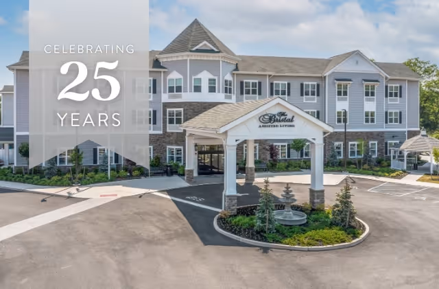 Front exterior of a three-story assisted living building with a covered porte-cochère, circular driveway and landscaping, with a translucent banner reading "CELEBRATING 25 YEARS".