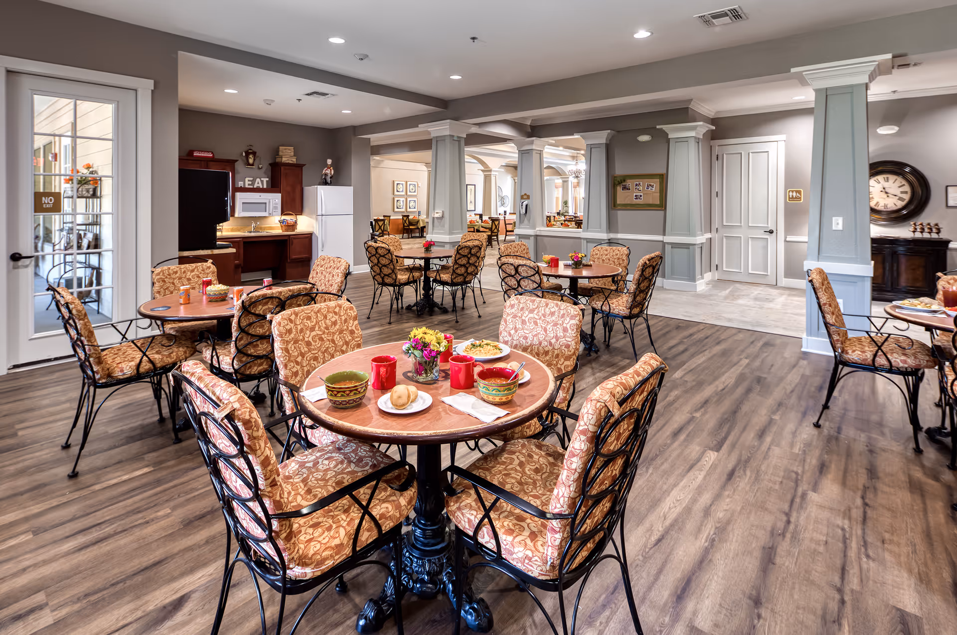 Bright communal dining room with round tables set for meals, upholstered chairs, and a service area in the background.
