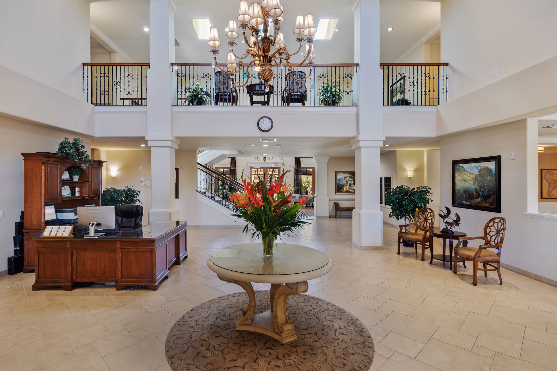 Spacious and elegant senior living facility lobby with a large round table holding a vase of red and orange flowers in the center. To the left, there is a wooden reception desk with a computer and chair. The area features beige tiled floors, white walls, and a high ceiling with a large chandelier. Upstairs balcony with black wrought iron railing overlooks the lobby. Seating area with two chairs and a small table is visible on the right side, along with framed artwork and potted plants.