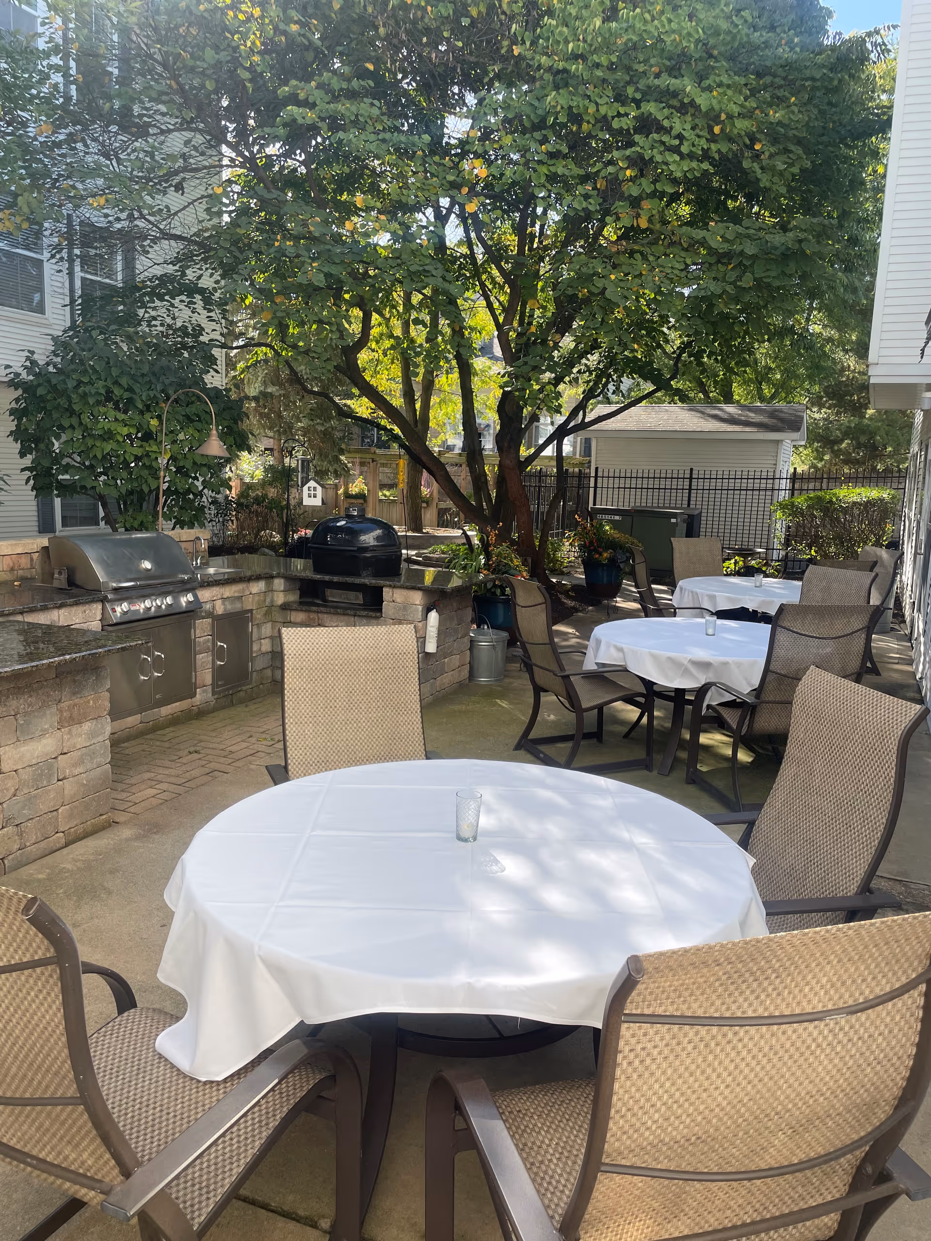 Outdoor patio with round tables draped in white tablecloths, several wicker chairs, a grill area, and trees providing shade.