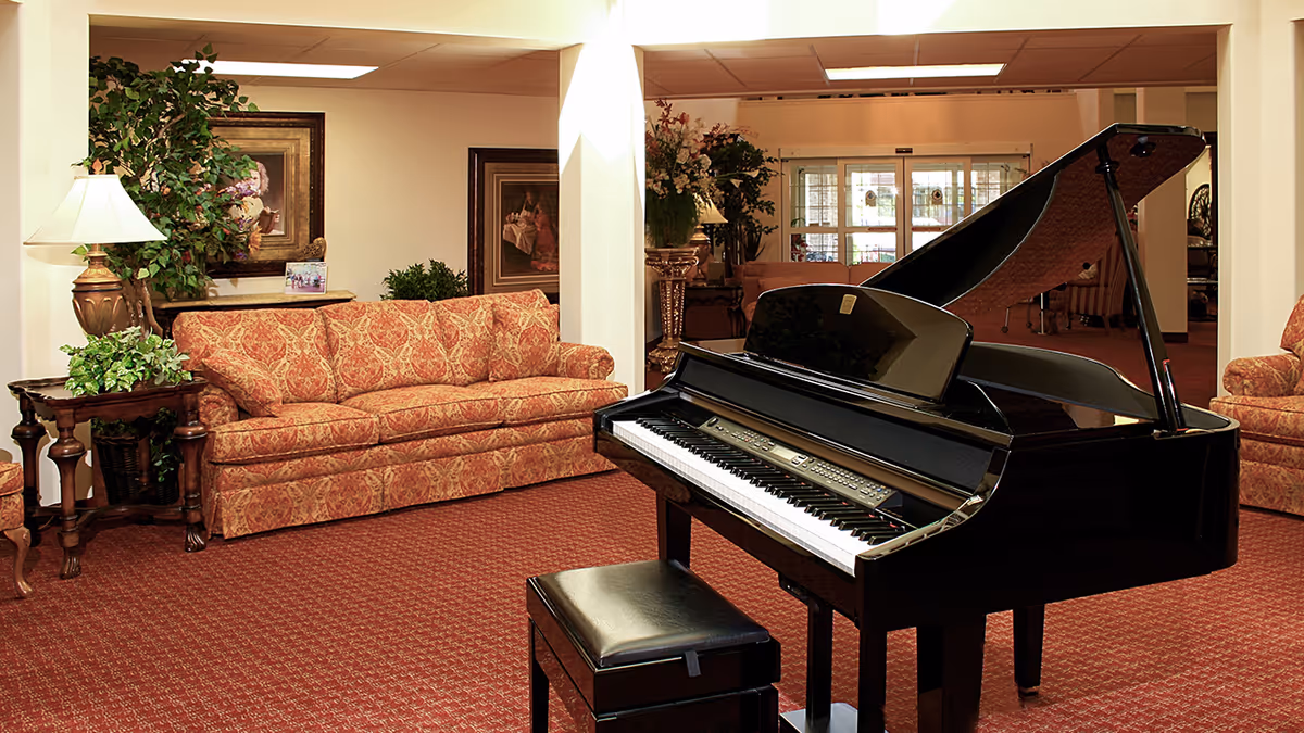 A carpeted lounge with a black grand piano, patterned sofas, side tables, lamps, and plants.