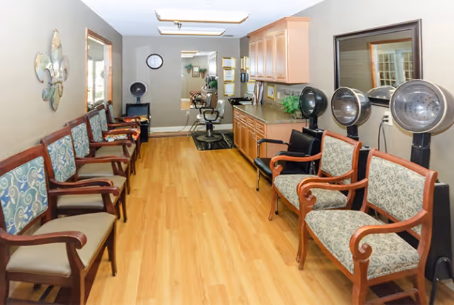 Interior view of a hair salon area in a senior living facility with wooden flooring, multiple chairs lined up on both sides, hair drying stations, a counter with cabinets, and mirrors on the walls.