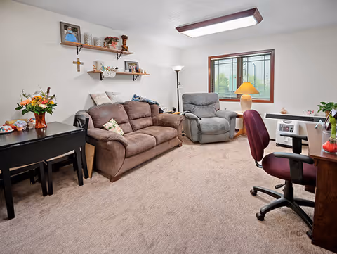 A cozy living room with a brown sofa, a gray recliner chair, and a desk with a maroon office chair. The room has beige carpet, a window with a view outside, a floor lamp, and shelves on the wall holding decorative items and framed photos.