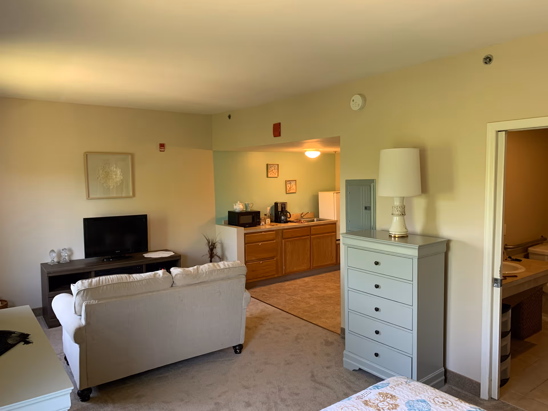Interior view of a living space in an assisted living facility showing a beige couch facing a TV on a wooden stand, a light blue chest of drawers with a lamp on top, an open doorway leading to a bathroom, and a small kitchen area with wooden cabinets, a microwave, coffee maker, and refrigerator.