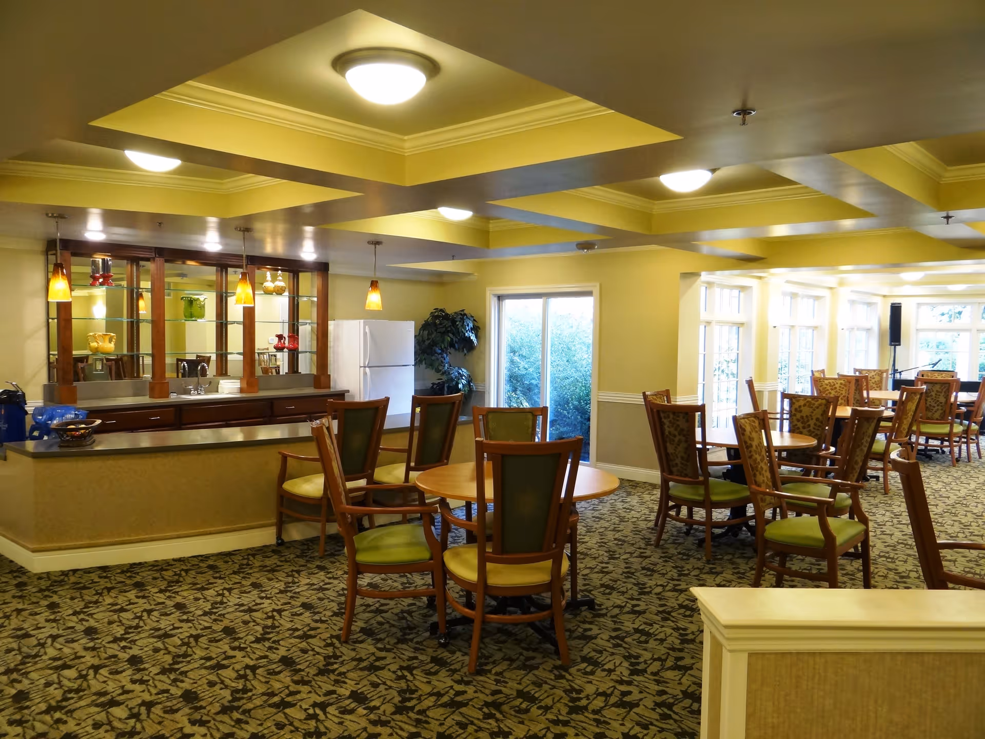 A spacious dining room area with multiple round wooden tables and chairs with green cushions. The room features a patterned carpet, a ceiling with recessed lighting, and large windows allowing natural light. There is a kitchen counter with a sink, hanging pendant lights, and a white refrigerator in the background.