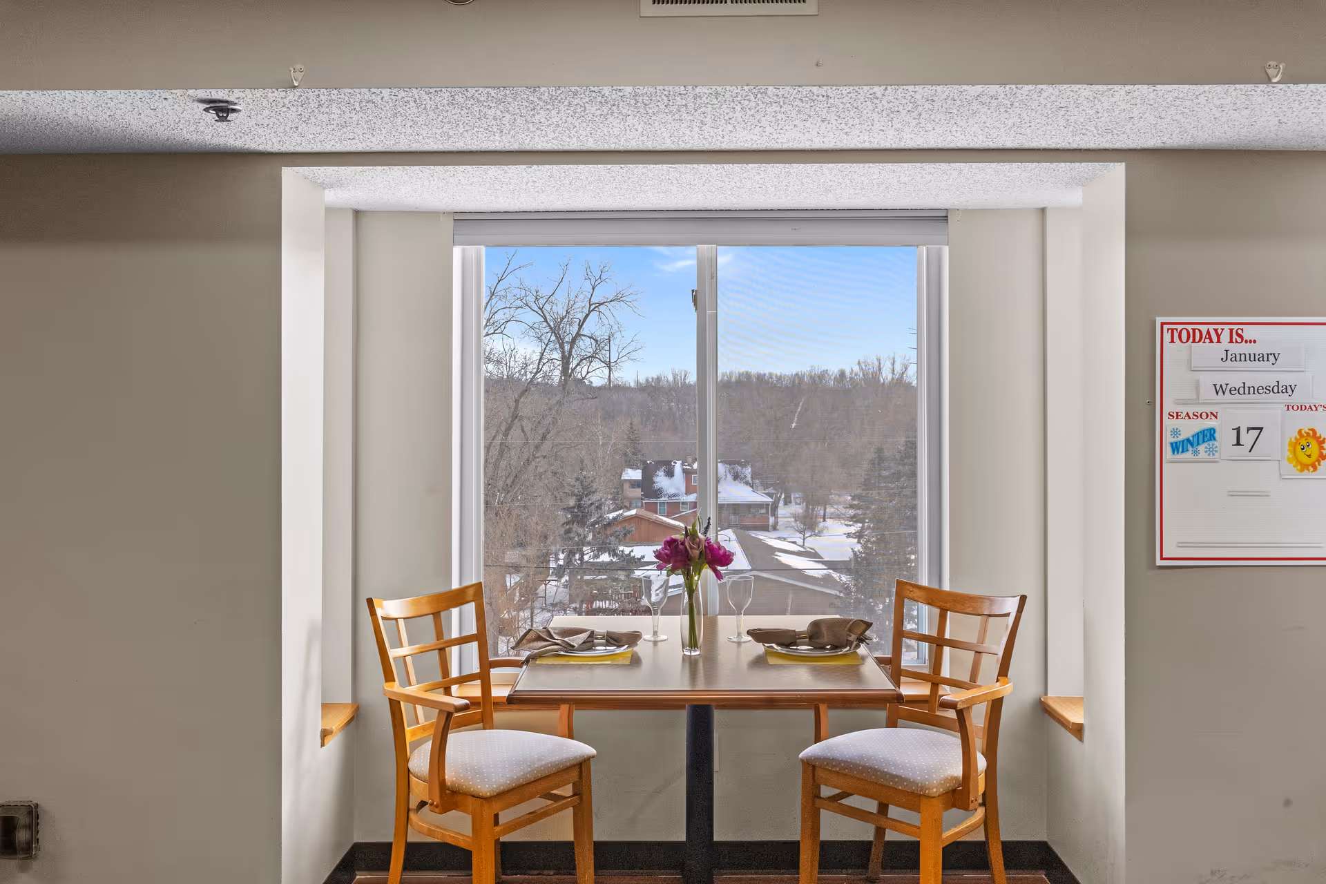 A small dining table set for two with wooden chairs and a vase of flowers in front of a large window showing a snowy outdoor scene with trees and rooftops. A whiteboard on the wall displays the date as Wednesday, January 17, with winter season and sunny weather icons.