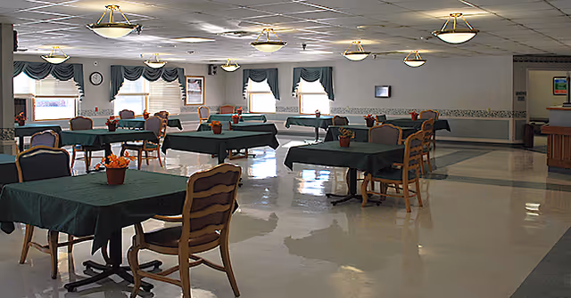 A spacious dining room with several tables covered in dark green tablecloths, each table decorated with a small flower arrangement. Wooden chairs with cushions surround the tables. The room has large windows with green valances and white blinds, and ceiling lights providing illumination. The floor is shiny and reflective.