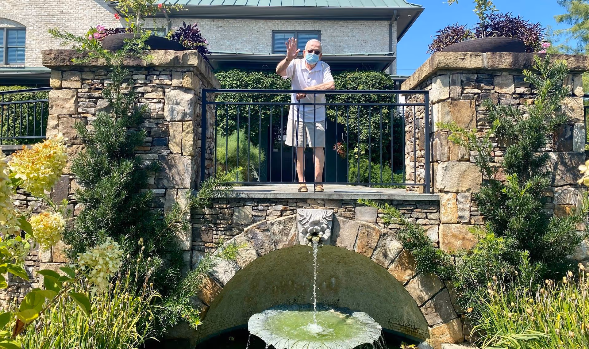 An elderly man wearing a face mask and casual summer clothes waves while standing on a stone balcony with a black metal railing. Below the balcony is a stone arch with a decorative water spout flowing into a small fountain surrounded by greenery and flowering plants. The background shows part of a building with beige brick walls and windows under a clear blue sky.