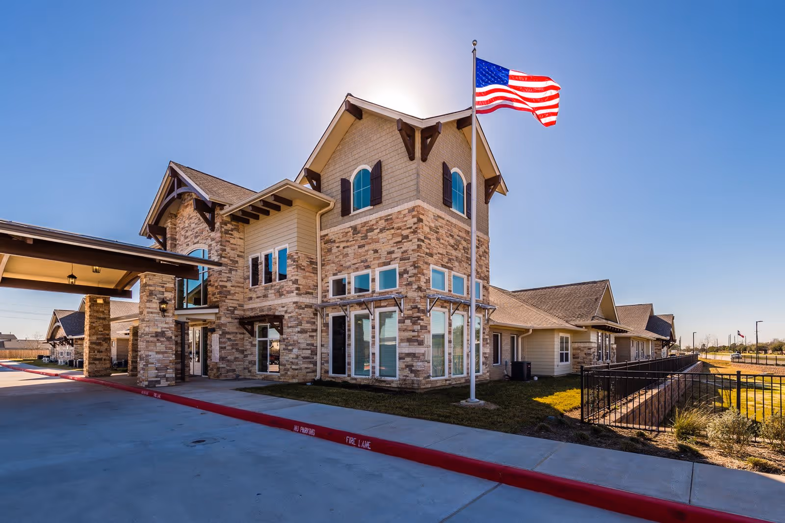 Exterior view of a senior living facility building with stone and beige siding, multiple windows, and a covered entrance. An American flag is flying on a flagpole in front of the building under a clear blue sky.