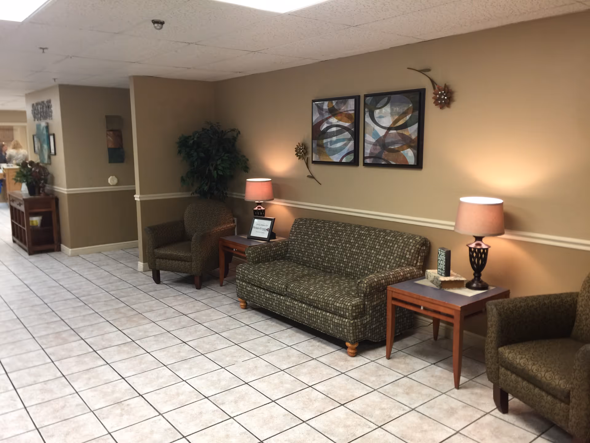 A hallway seating area in a senior living facility with two armchairs and a patterned sofa arranged around two wooden side tables with lamps. The walls are beige with two abstract paintings and decorative wall accents. The floor is tiled with white and gray tiles, and there is a potted plant in the corner.