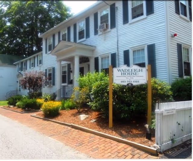Exterior view of Wadleigh House Assisted Living, a two-story white building with black shutters and a small porch with columns. There is a garden with bushes and flowers in front, and a sign displaying the facility's name and phone number.