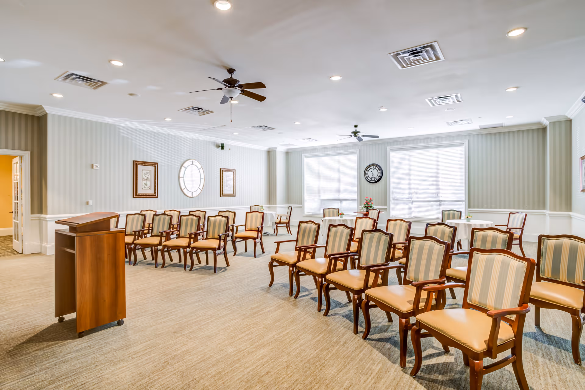 A bright, spacious room with rows of wooden chairs featuring striped upholstery arranged facing a wooden podium. The room has light-colored walls with decorative molding, two large windows with blinds, ceiling fans, recessed lighting, and framed artwork on the walls.