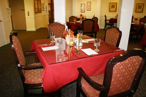 Dining room with tables covered in red tablecloths, set with glasses, napkins, and condiments. Upholstered chairs with patterned fabric surround the tables. The room has carpeted floors, framed artwork on the walls, and soft lighting.
