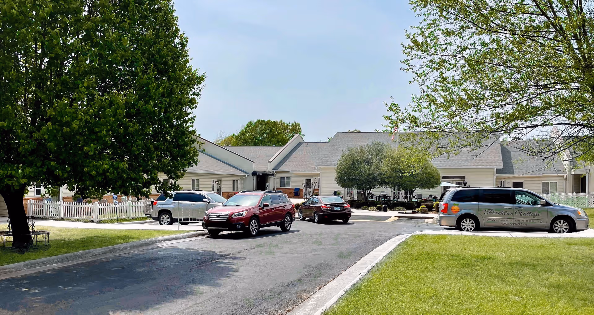 Exterior view of Peachtree Village Assisted Living of Fort Smith, showing a single-story building with a gray roof, surrounded by trees and a grassy area. Several cars are parked in front, including a van with the facility's name and contact information on the side.