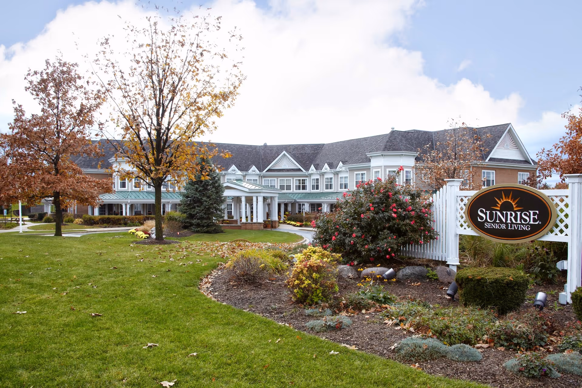 Exterior view of Sunrise Senior Living facility in Palos Park, showing a large two-story building with white trim and a covered entrance. The foreground features a well-maintained lawn, trees with autumn leaves, and landscaped garden beds. A white fence with a sign reading 'Sunrise Senior Living' is visible on the right side.