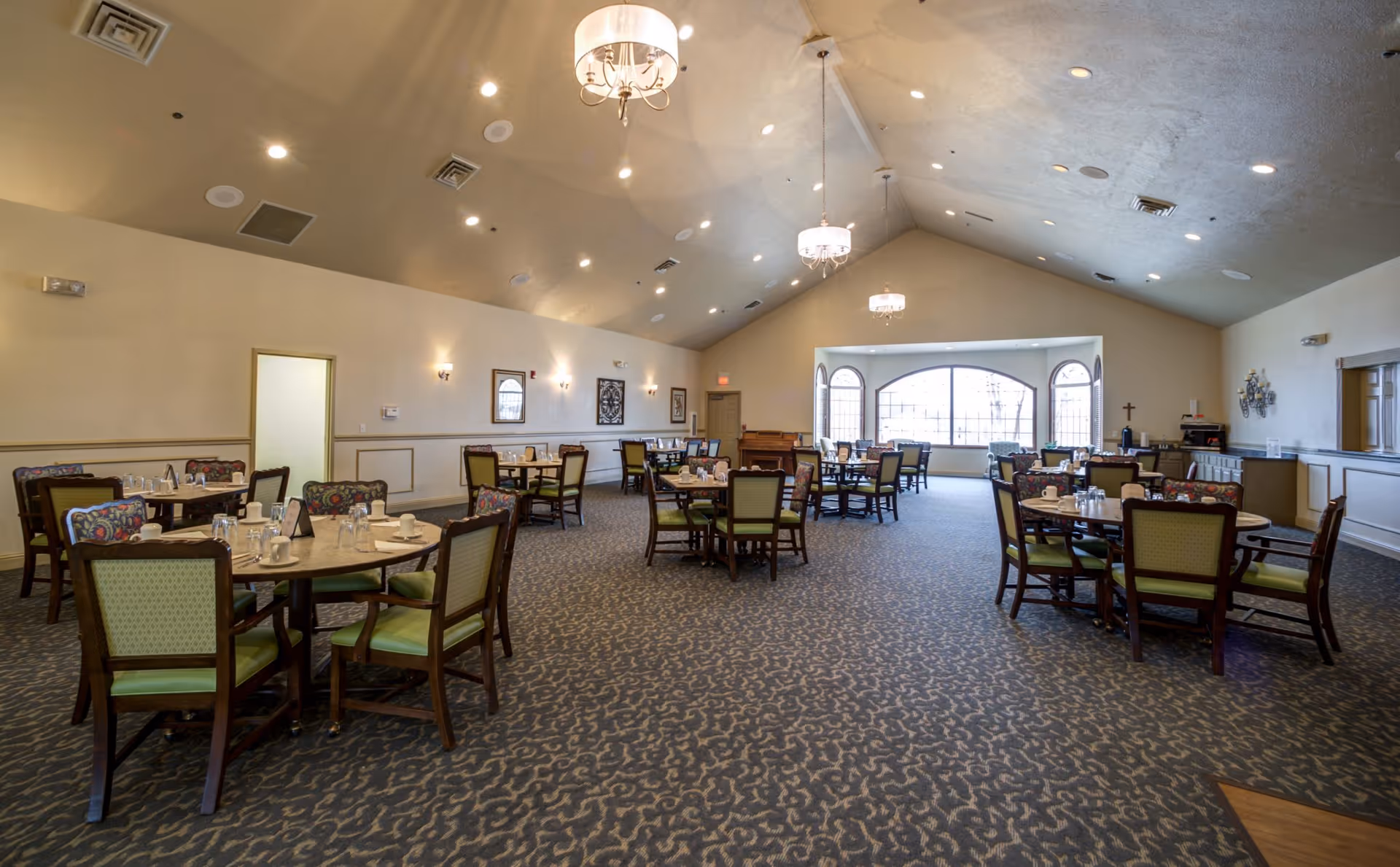 Spacious dining room with multiple round tables and green-upholstered chairs under chandeliers and a large arched window.