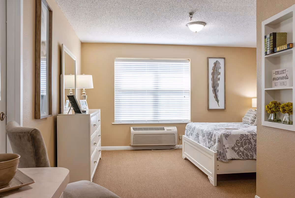 A cozy bedroom with beige walls and carpet, featuring a white bed with patterned bedding, a white dresser with a lamp and framed photos, a window with blinds, and a built-in wall shelf holding books, decorative items, and a sign that reads 'Collect moments not things'.