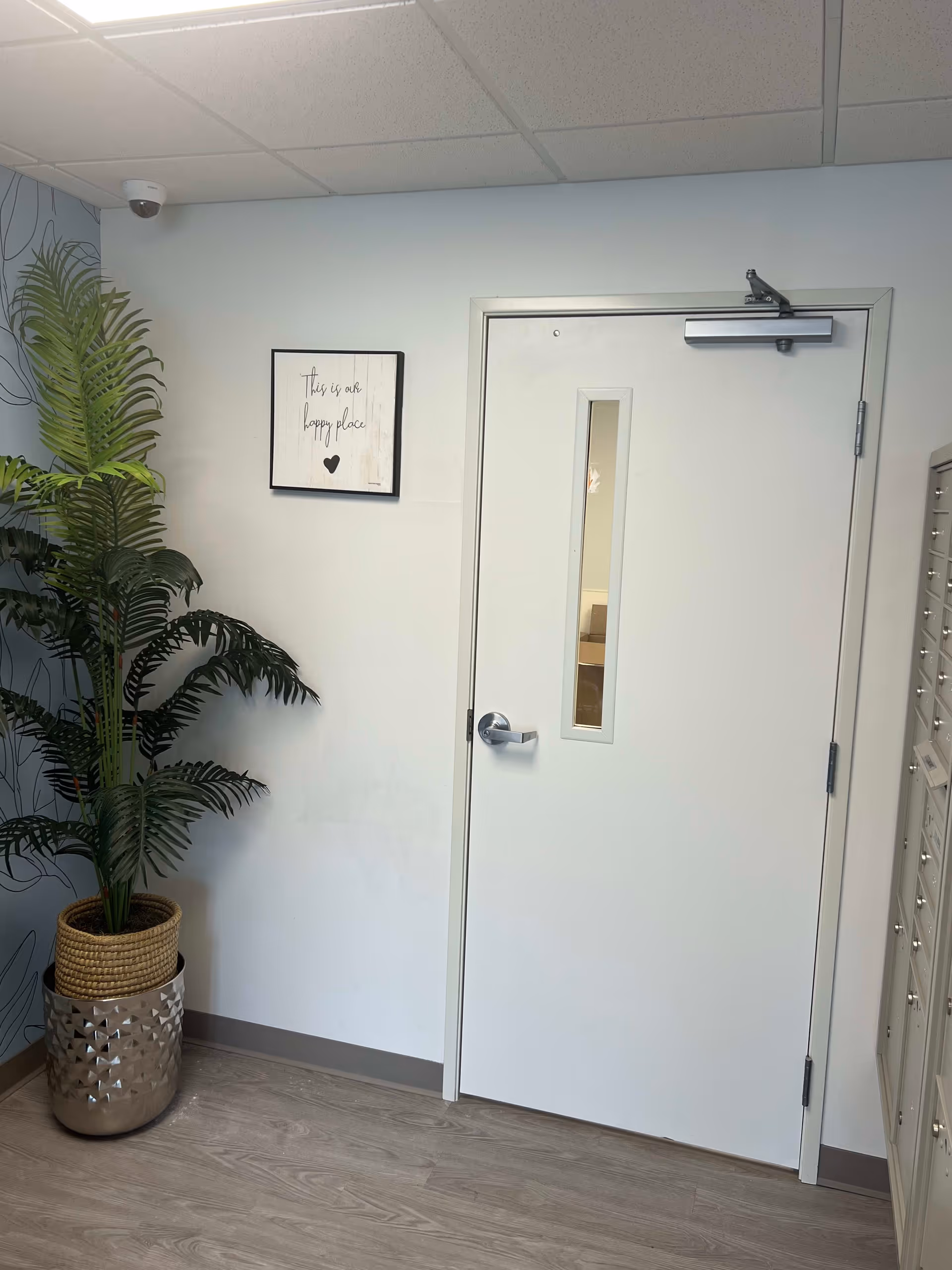 Interior corner of a room with a white door featuring a narrow vertical window. To the left of the door is a potted artificial plant with large green leaves in a decorative pot. Above the plant on the wall is a framed sign that reads 'This is our happy place' with a small heart below the text. The floor is wood-patterned, and there is a security camera mounted on the ceiling in the corner.