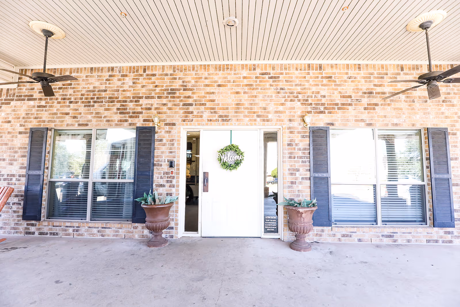 Front entrance of a building with a white door decorated with a green wreath that says 'Welcome'. The entrance is flanked by two large windows with black shutters and two large brown planters with green plants. The ceiling above the entrance has two black ceiling fans.
