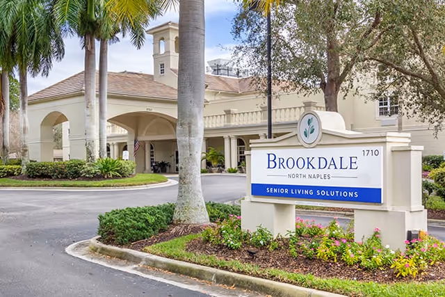 Exterior view of Brookdale North Naples senior living facility showing the entrance with a covered driveway, palm trees, and a sign that reads 'Brookdale North Naples Senior Living Solutions'.