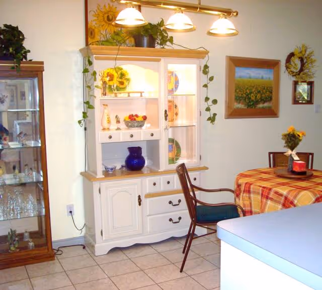 A cozy dining area featuring a white wooden hutch with glass doors displaying decorative plates and vases, a wooden chair with a blue cushion, a round table covered with a red and yellow plaid tablecloth, a small flower arrangement on the table, and framed sunflower-themed artwork on the wall.