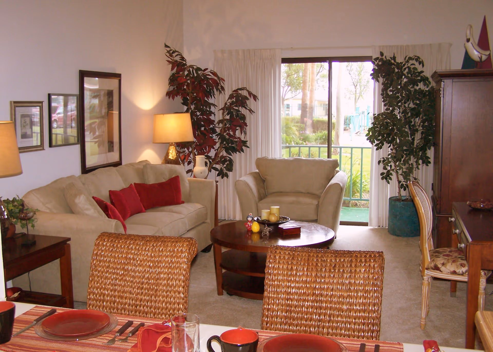 A cozy living room with a beige sofa and armchair, both adorned with red cushions. There is a round wooden coffee table in the center with decorative items on it. Two large potted plants flank a sliding glass door that opens to a green outdoor area. A wooden desk with a floral cushioned chair is on the right side, and a dining table with woven chairs and red table settings is partially visible in the foreground.