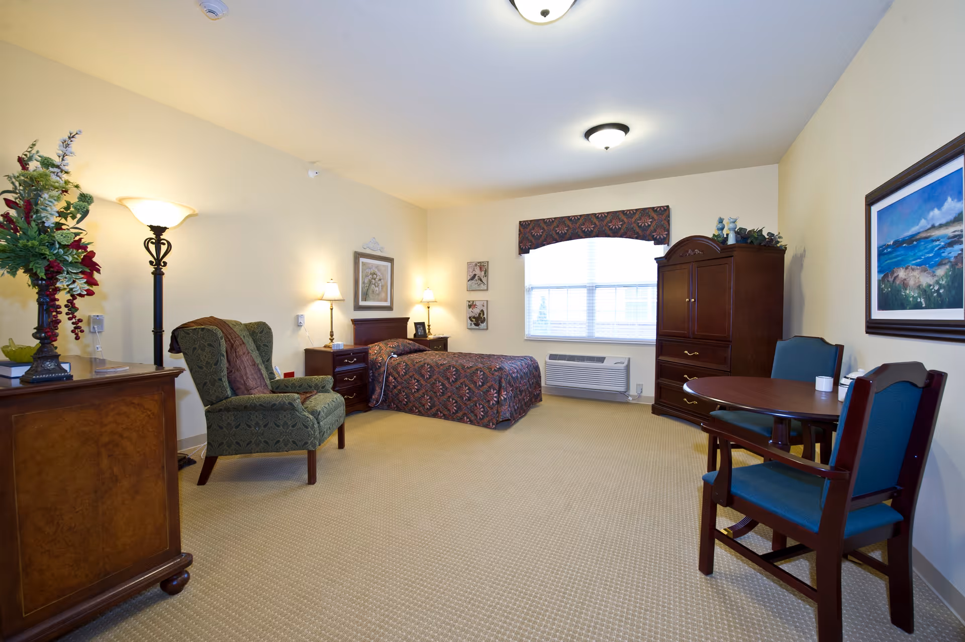 A well-lit senior living bedroom with a single bed covered in a patterned bedspread, two nightstands with lamps, a green upholstered armchair, a wooden dresser with a floral arrangement, a wooden armoire, and a small round table with two chairs. There is a large window with blinds and a decorative valance, beige carpet, and framed artwork on the walls.