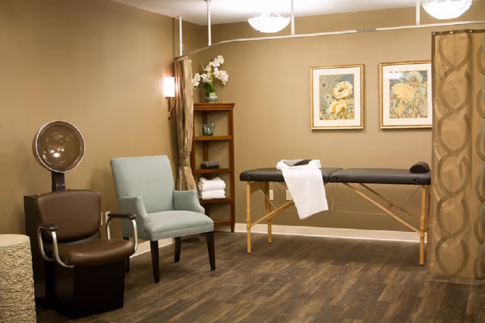 A cozy room with a massage table covered with a white towel, two chairs including a brown salon chair and a light blue armchair, a wooden shelf with folded towels and decorative items, and two framed floral paintings on the wall. The room has warm beige walls and wood flooring.