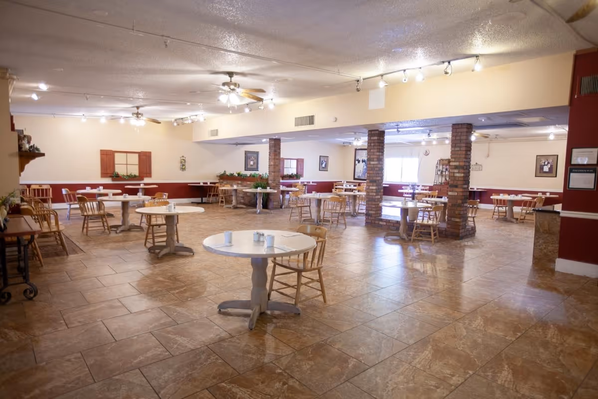 Spacious dining room with multiple round tables and wooden chairs arranged neatly. The room has tiled floors, brick pillars, ceiling fans with lights, and soft yellow walls with red accents. Some tables have cups and small items on them, and there are framed pictures and decorative plants on the walls.