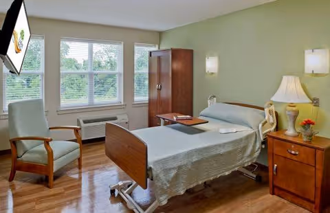 A clean and well-lit hospital-style bedroom with a single adjustable bed covered with a light gray blanket, a wooden nightstand with a lamp and a small vase of flowers, a wooden wardrobe, a comfortable armchair, and three large windows with white blinds letting in natural light.