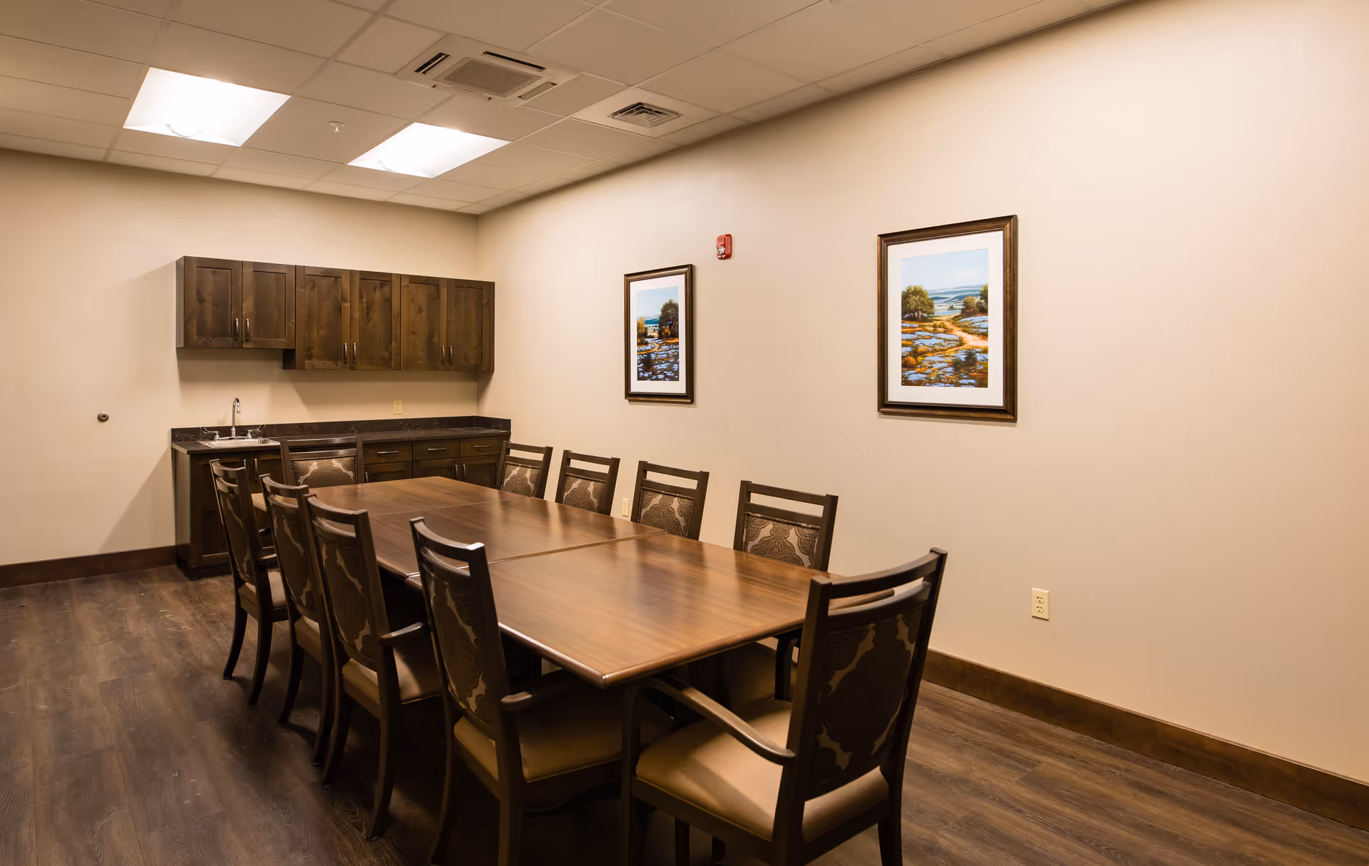Long wooden dining table surrounded by chairs in a room with a small kitchenette, wood cabinets, and framed landscape paintings on the wall.