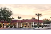 Single-story beige nursing facility with a covered entrance, columns, palm trees and a parking area under a pastel sky.