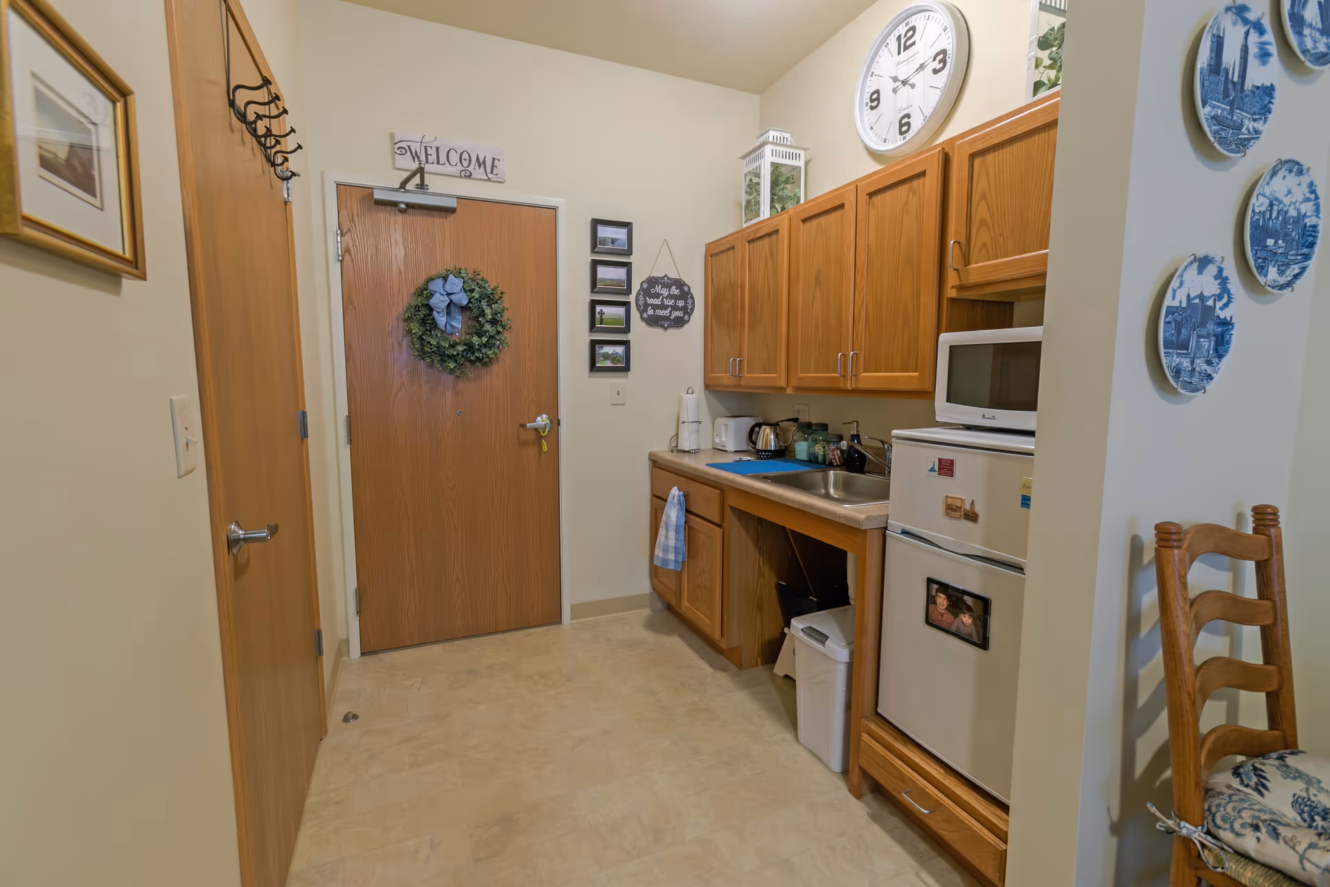 A small kitchen area with wooden cabinets, a sink, a microwave on top of a mini refrigerator, and a countertop with a kettle and other small items. A wooden door with a green wreath and a 'Welcome' sign above it is visible. There are decorative plates on the wall and a wooden chair with a cushion in the foreground.
