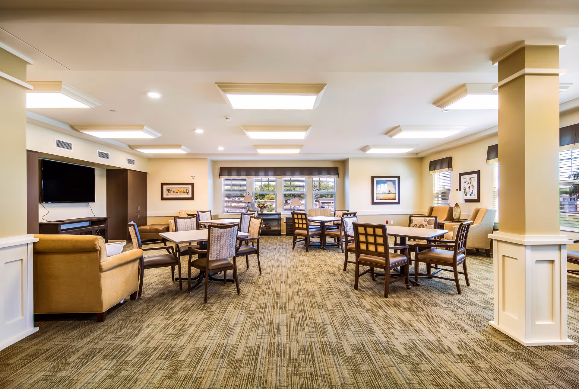 A bright and spacious common area with multiple tables and chairs arranged for seating. The room features beige walls, carpeted flooring, large windows with blinds, framed artwork on the walls, and a flat-screen TV mounted on a dark wood cabinet. There are also comfortable armchairs and lamps near the windows.