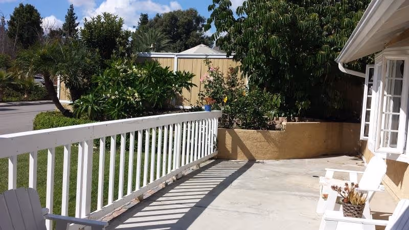 Outdoor patio area with white wooden railing and white chairs, potted plants, and greenery including trees and bushes under a blue sky with some clouds.