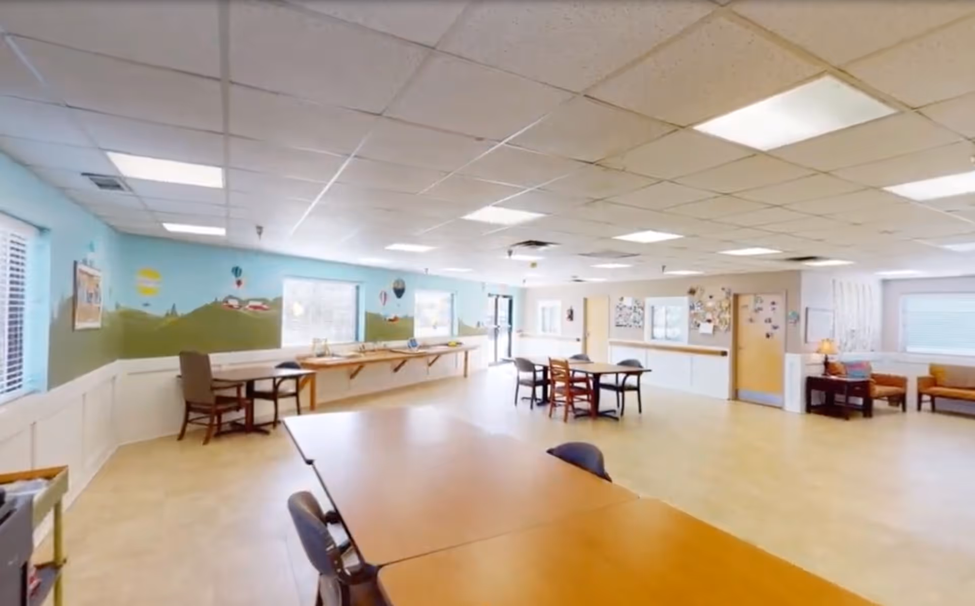 A spacious, well-lit common area with several tables and chairs arranged for group activities or dining. The walls are painted light blue with a mural of green hills, houses, and hot air balloons. Large windows allow natural light to fill the room, and there are bulletin boards and seating areas along the walls.