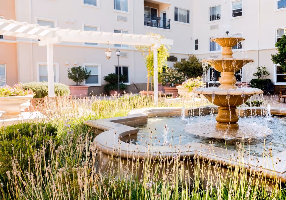 Outdoor courtyard area of Heritage Estate Senior Apartments featuring a multi-tiered stone fountain with water flowing, surrounded by plants and flowers. The background shows the exterior walls and windows of the apartment building with a white pergola and hanging lanterns.
