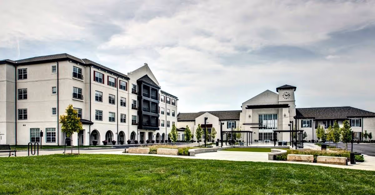 Exterior view of a large, modern senior living facility named Norterre, featuring a multi-story building with numerous windows, a clock tower, landscaped green lawn, benches, and paved walkways under a partly cloudy sky.