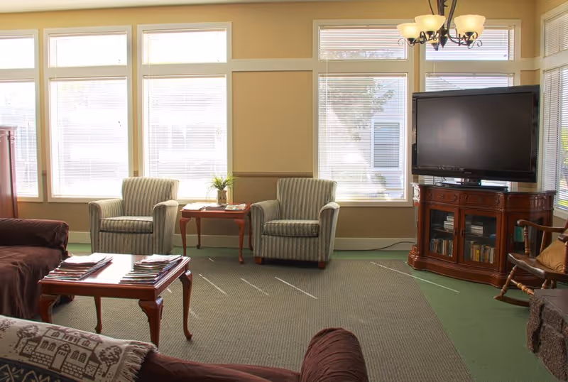 A well-lit living room with large windows covered by blinds. The room features two striped armchairs with a small wooden table between them, a wooden TV stand with a flat-screen television, a rocking chair, and two brown sofas. A wooden coffee table with magazines is in the center of the room, and the floor is carpeted in green and beige tones.